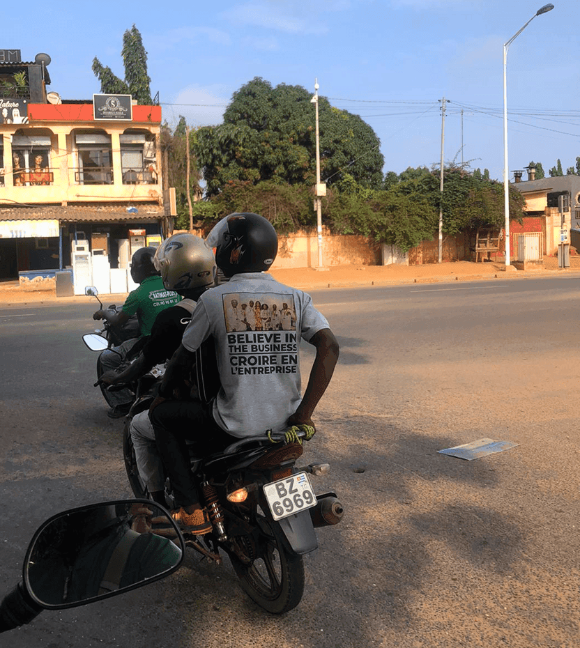 Two men on a scooter in a street in Togo. On the T-shirt of the man in the back, we can read “Believe in the Business” written in English and French.