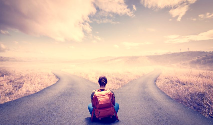 Woman with a backpack on sitting contemplatively at the fork of a road that has no signs to indicate direction