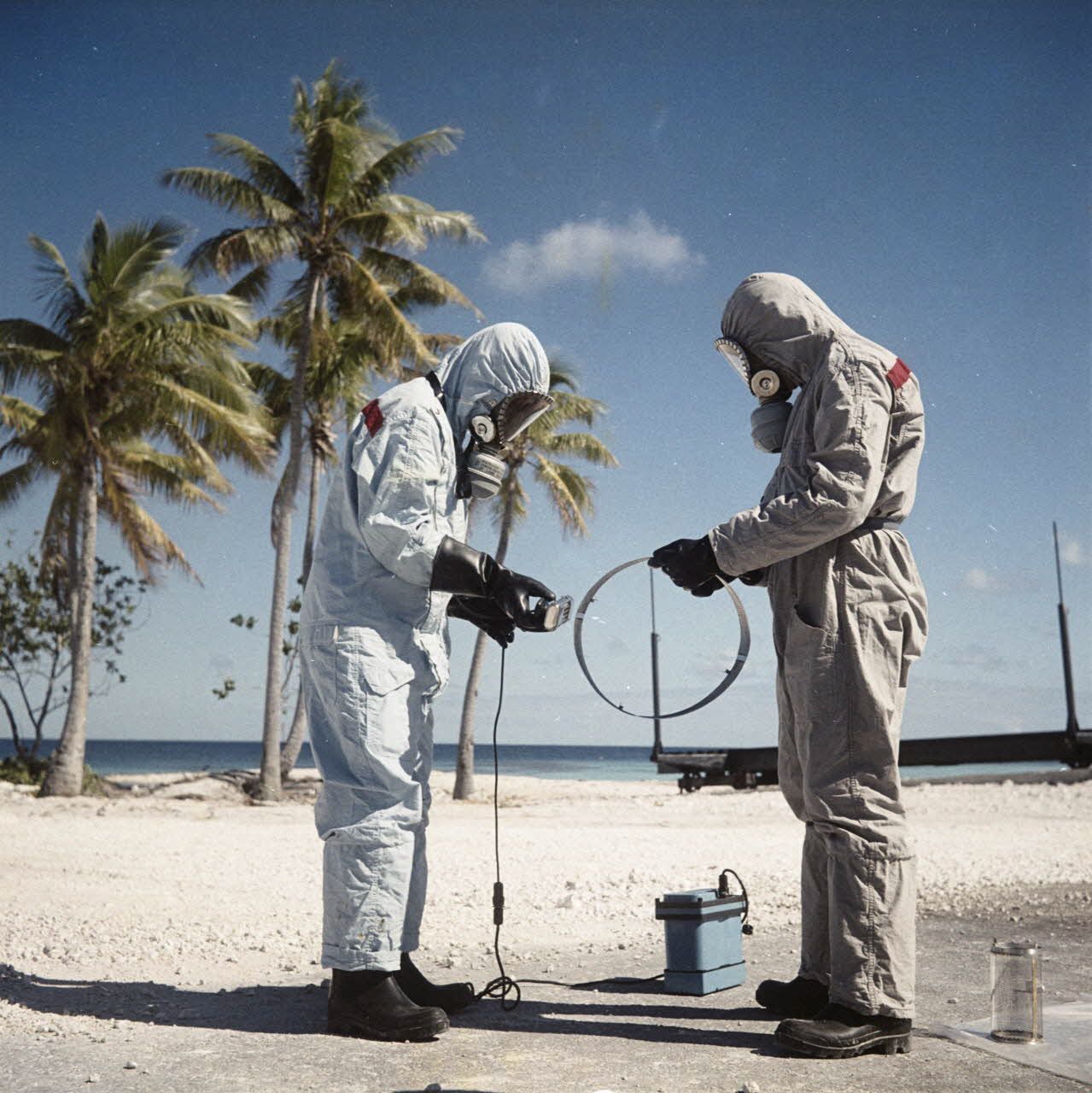 Sur une aire du service de décontamination de Hao, des personnels en tenue de protection testent la radioactivité de matériel avec un détecteur après un tir d'essai nucléaire (juillet 1966) - © Robert Antoine/ECPAD/Défense