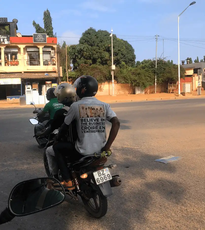 Two men on a scooter in a street in Togo. On the T-shirt of the man in the back, we can read “Believe in the Business” written in English and French.