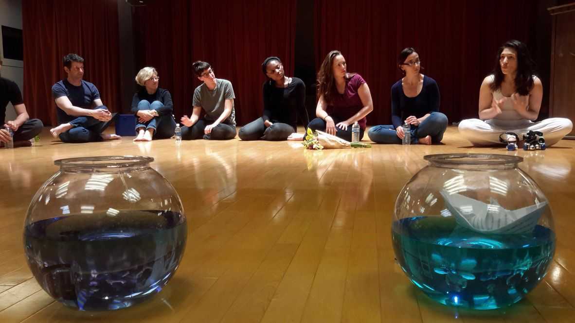 Huit personnes assises en demi-cercle sur le parquet d'une salle de théâtre, devant des rideaux rouges, en pleine discussion. Au premier plan, deux bocaux en verre remplis d'eau bleue, dont l'un contient un bateau en papier.